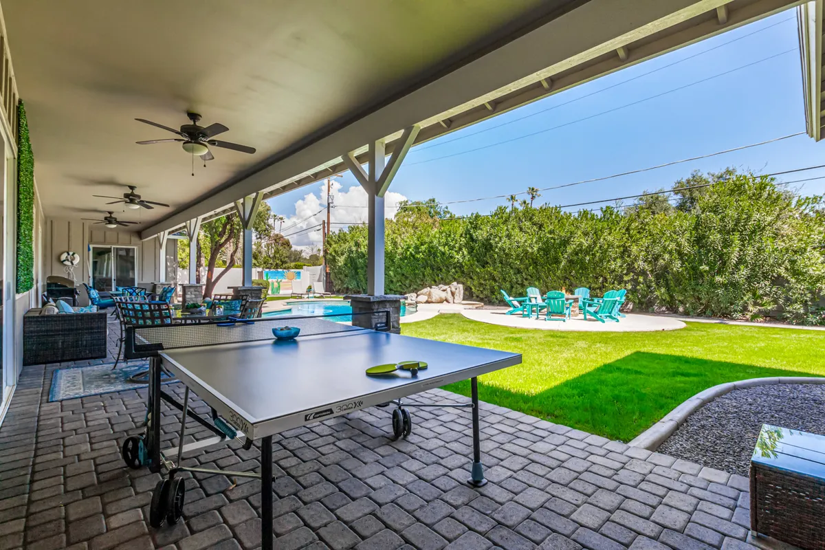 A lively outdoor covered patio space with a ping pong table as the focal point, surrounded by a swimming pool, lounge seating, and a green lawn. The scene brims with possibilities for outdoor games and group activities.