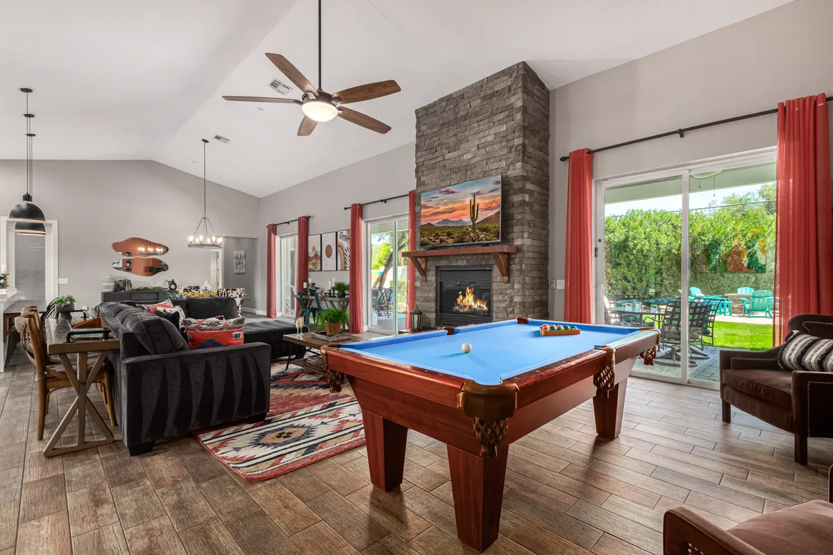 This image captures the open-plan living area featuring a pool table as a centerpiece, a comfortable sectional sofa, a cozy fireplace, and large windows that flood the space with natural light. Decorative artwork adds a stylish touch, and the room feels inviting and perfect for socializing or relaxing.
