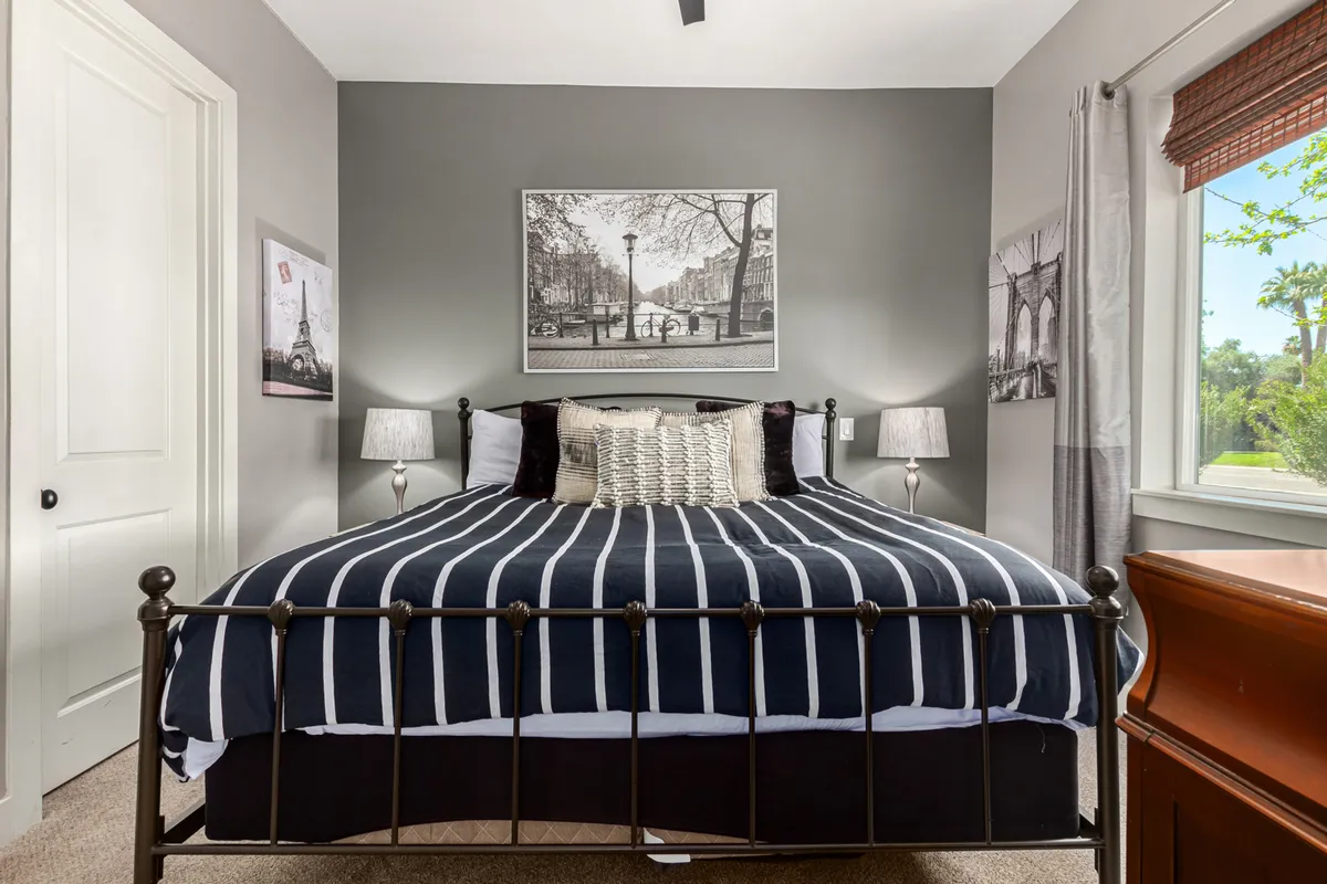 A serene bedroom featuring a large bed with striped bedding, black and white wall art, matching nightstands with lamps, and a wooden dresser. The room is bathed in natural light from a nearby window, creating a soothing and tranquil ambiance ideal for restful sleep.