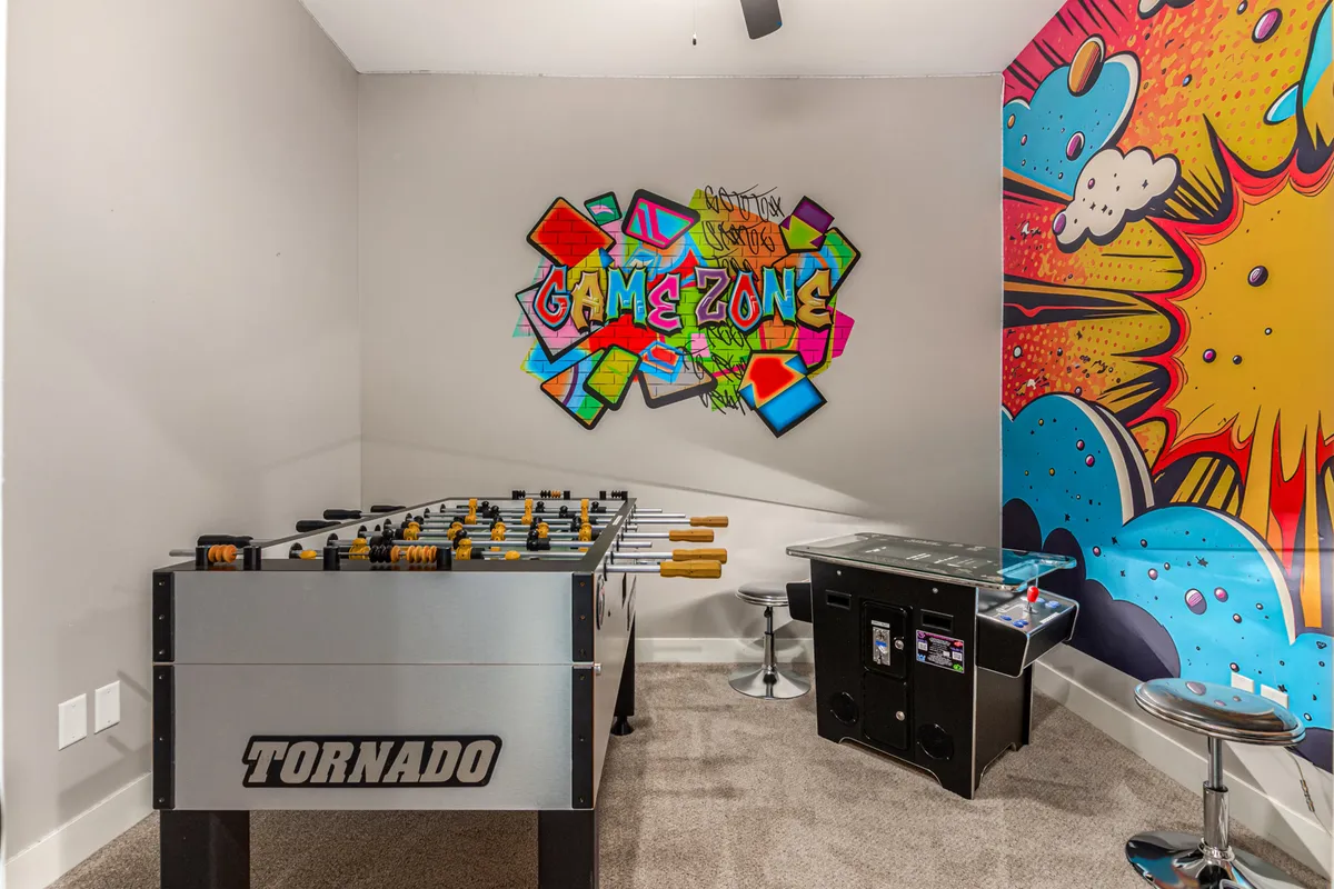 A vibrant game room featuring a foosball table, a classic arcade game cabinet, and distinctive graffiti-inspired wall art. The lively atmosphere is enhanced by colorful decor and casual seating, instantly evoking the nostalgia of an old-school arcade.