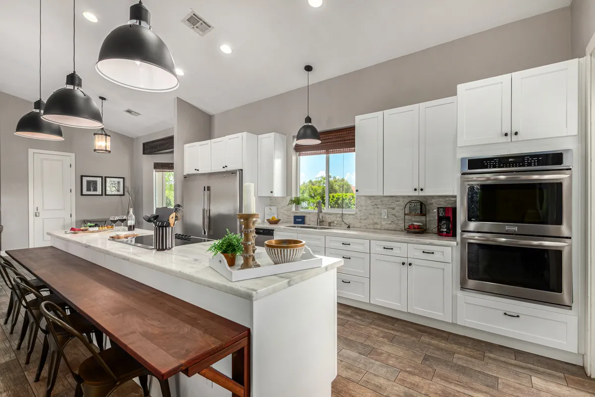 The image showcases a modern, gourmet kitchen with a large island, stainless steel appliances, and a dining table nearby. Ample natural light streams in through windows, creating a warm, welcoming ambiance ideal for cooking, dining, and socializing.