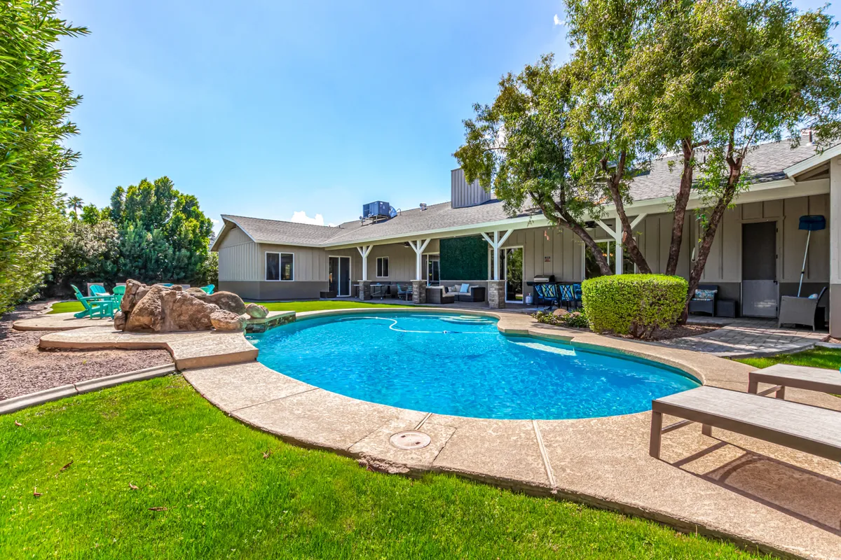 The hero image showcases a sun-drenched backyard featuring a sparkling heated swimming pool, elegant stone accents, inviting lounge chairs surrounded by lush green hedges, and a spacious patio area perfect for gatherings. The scene radiates relaxation and leisure, highlighting spaces for swimming, sunbathing, and outdoor dining—all set against the backdrop of clear Scottsdale skies.