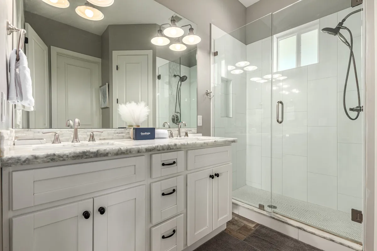 A luxurious bathroom with a double sink vanity, a glass shower enclosure, a marble countertop, and a large mirror with lighting fixtures. The space exudes a spa-like atmosphere perfect for morning routines or unwinding after a long day.